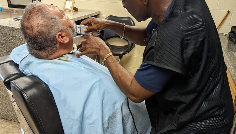 A student barber using electric trimmers to groom a customer's beard in a barber college.