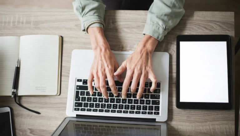 A copywriter typing on a laptop computer with a digital tablet and notebook on a desk.