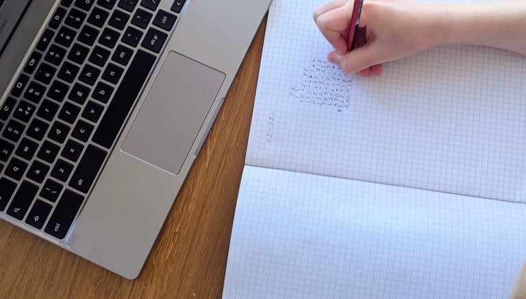 An 11+ student's hand writing math in a grid notebook next to a silver laptop learning at home