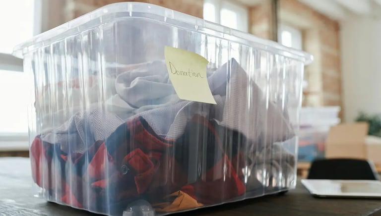 A clear plastic storage bin filled with folded clothes for donation labeled with a sticky note.