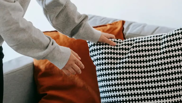 A person in a gray hoodie arranging patterned and solid orange throw pillows on a modern gray sofa.