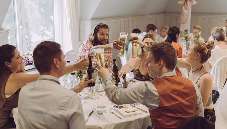 Hochzeitsgäste stoßen fröhlich mit Biergläsern am Tisch an. Authentische Eventfotografie von Just Perspectives.