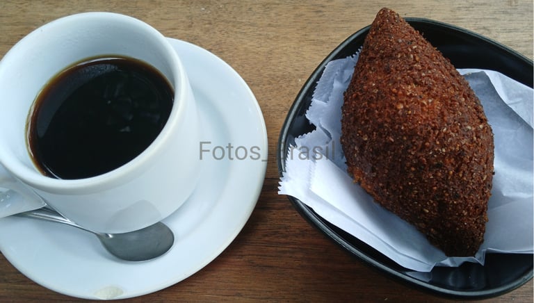 Uma xícara de café fumegante sobre um pires branco, ao lado num pratinho, um kibe dourado e crocante