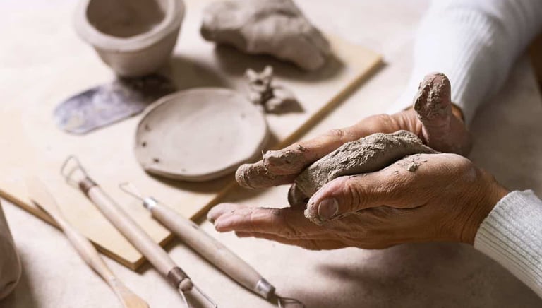 A pair of hands mold a piece of clay on a work surface, surrounded by various pottery tools and partially shaped clay objects