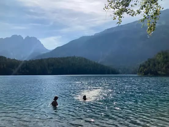 Couple swimming in calm alpine lake with sunlit mountain backdrop and forested hills in Tyrol