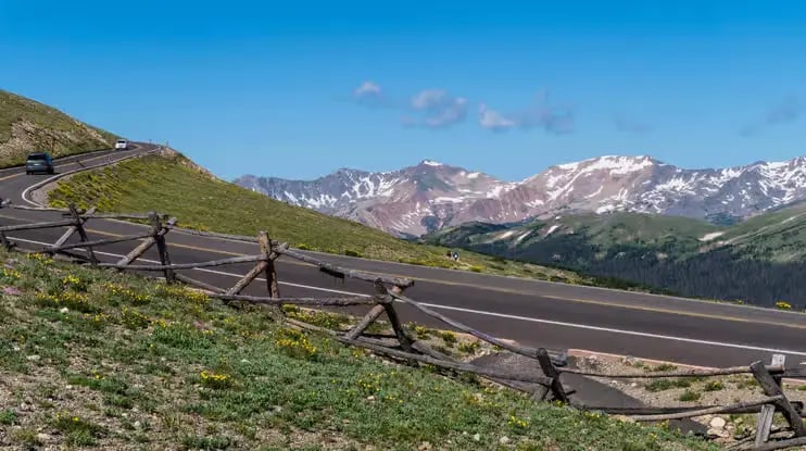 Winding asphalt road with wooden fences overlooking snow-capped Rocky Mountains.