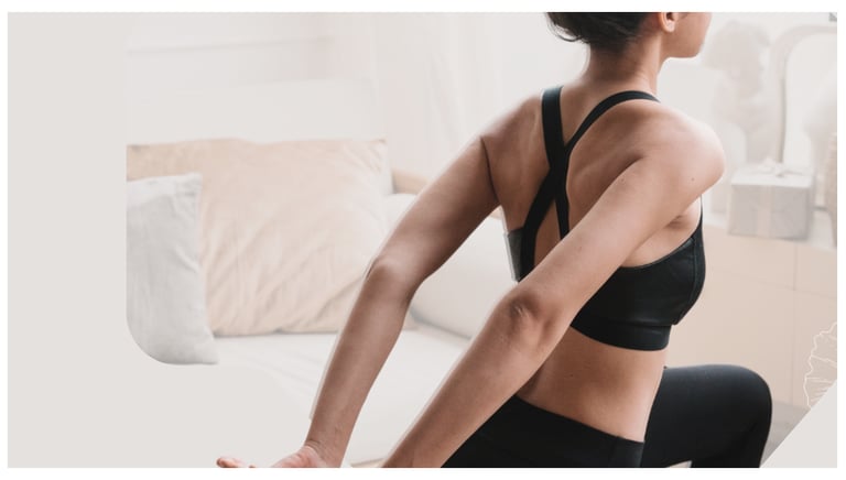 A woman in a black sports bra performing a chest and shoulder stretch in a bright living room.
