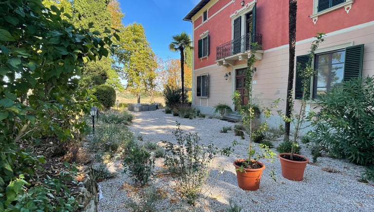 Villa Violetta with a red facade and gravel courtyard surrounded by lush Mediterranean plants.