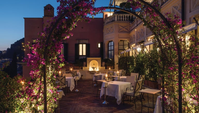 Luxury terrace restaurant in Italy at night with white tablecloths and a purple bougainvillea flower arch.