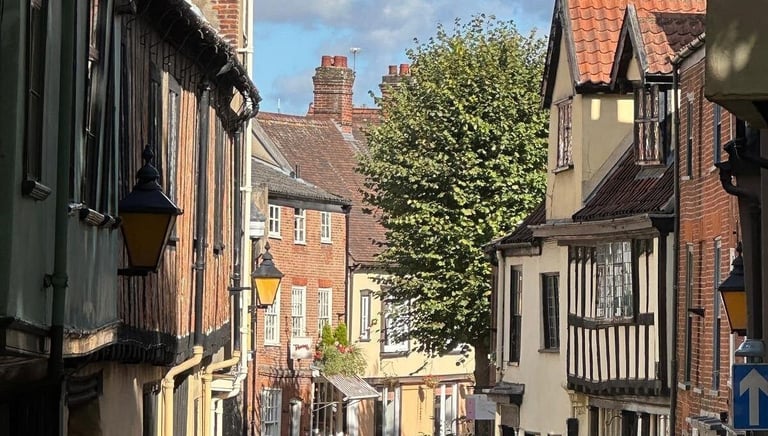 Traditional half-timbered buildings and brick houses lining a narrow historic street in a charming English village.