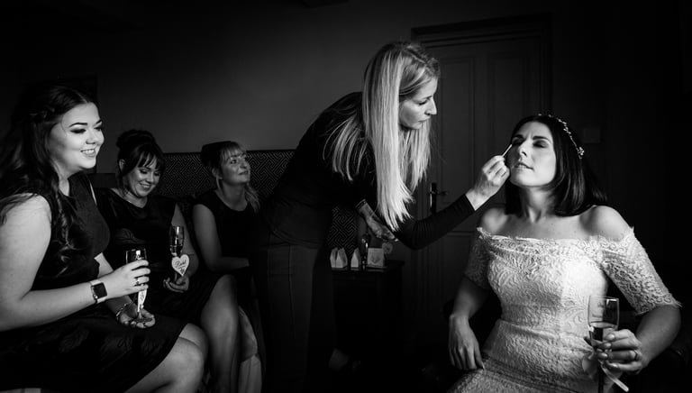 Professional makeup artist applying eye makeup to a bride in a lace wedding dress with bridesmaids.
