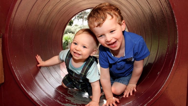 male toddler and older brother playing in play tunnel at a playground 