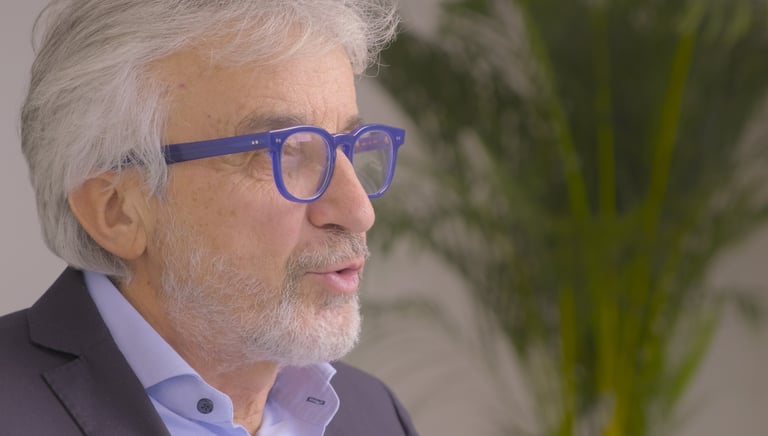 A senior professional man with grey hair and blue glasses speaking in a modern office.