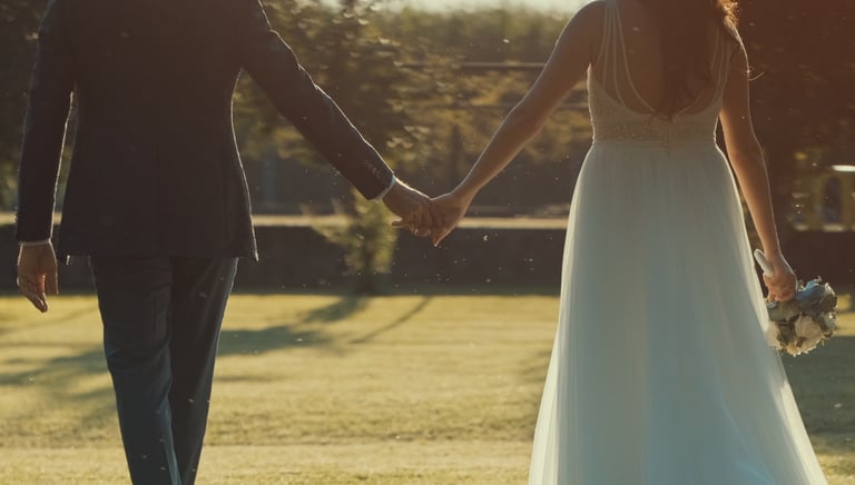 A bride and groom holding hands while walking through a sunny garden during an outdoor wedding ceremony.