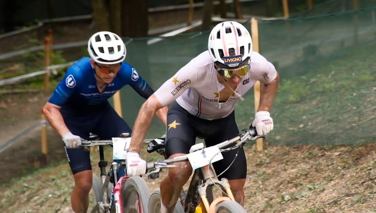Two professional cross-country mountain bikers racing uphill on a dirt trail wearing helmets and cycling kits.