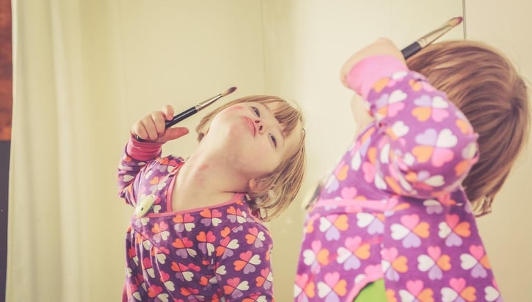 Young girl with Down syndrome playing with a makeup brush in front of a mirror.
