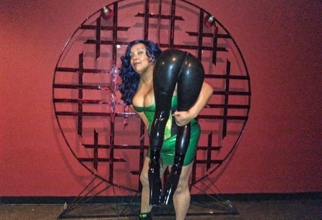 MsMadyson in a green latex dress carrying  a person over her shoulder who is wearing latex pants