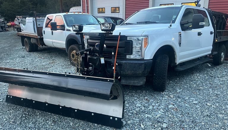 two white trucks parked in front of a shop. One truck has a plough blade and salter on it