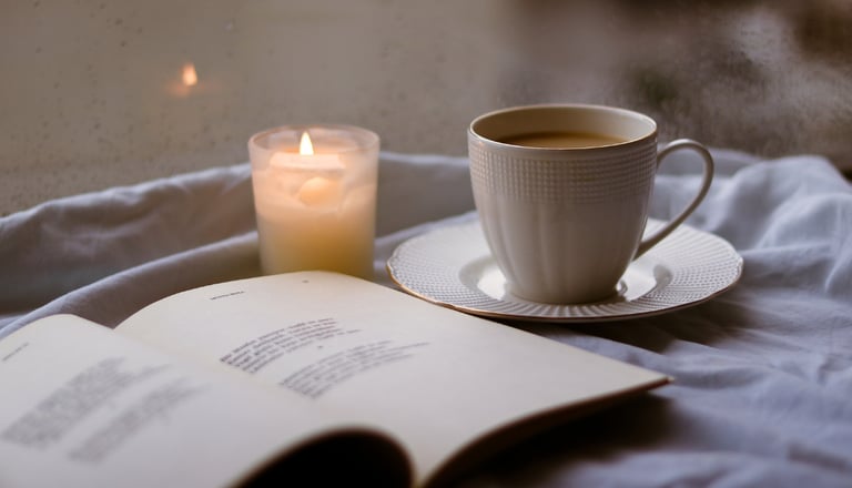 A cozy scene featuring an open book, a cup of hot coffee, and a lit candle on a bed.