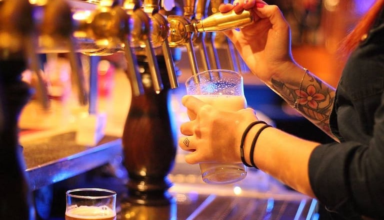A bartender with tattoos pouring a fresh craft beer from a brass tap into a glass at a pub.