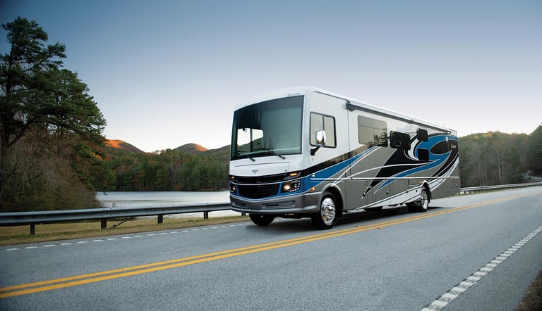 A luxury Class A motorhome driving along a scenic mountain road beside a peaceful lake at sunset.
