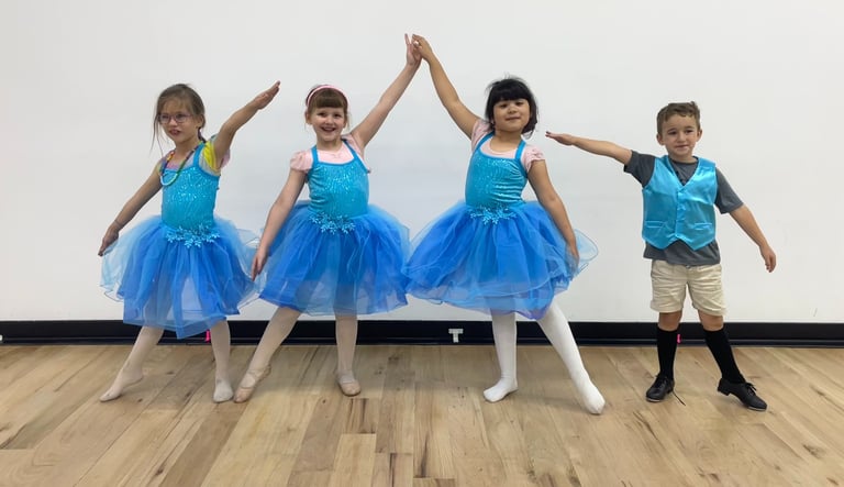 Young children in blue ballet tutus and dance costumes posing in a dance studio with wood floors.