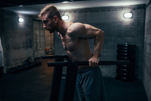 Muscular man performing triceps dips on parallel bars in a dark industrial gym setting.