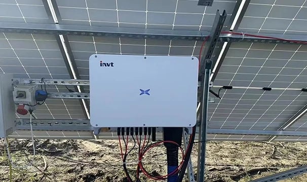 An INVT solar inverter mounted on a metal rack beneath ground-level solar panels.