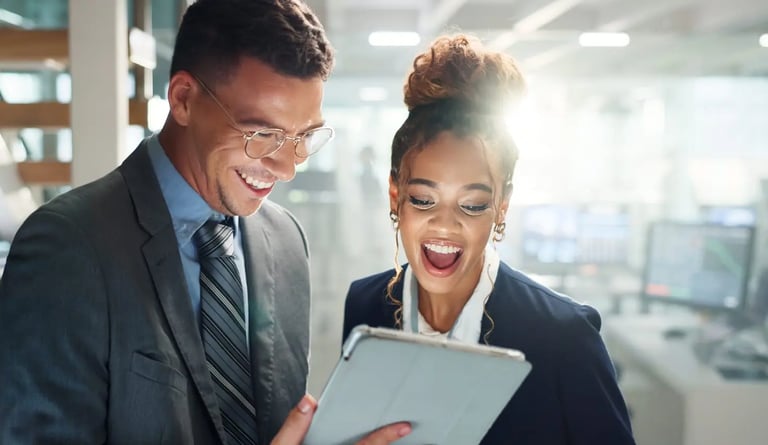 Two professionals stand close together in an office, smiling and looking at a tablet.