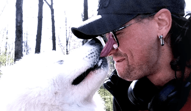 a photo of a man wearing a black baseball cap and glasses being licked in the face by white dog
