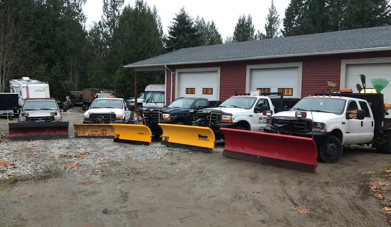 5 trucks with plows are parked in front of a 3 bay shop. The trucks are in a semi circle facing the camera