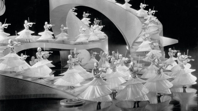 Female dancers in circular costumes playing white violins on a black‑and‑white Busby Berkeley set.