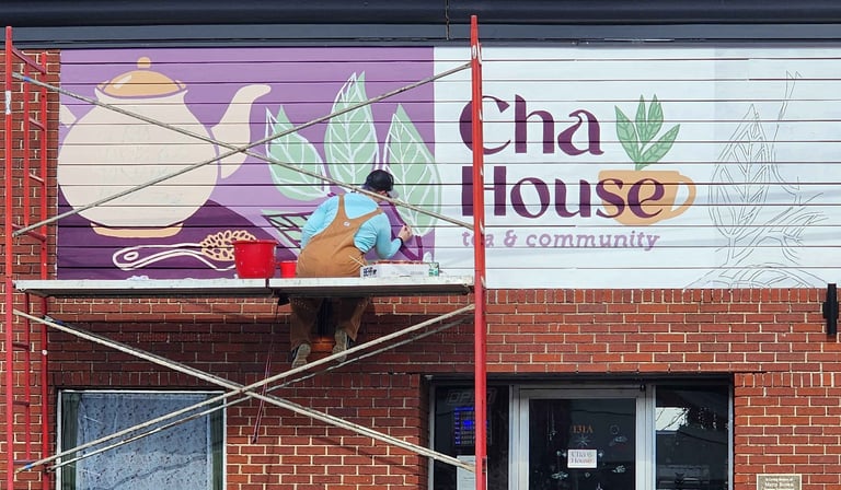 Artist on scaffolding painting a tea house mural with teapot and leaf graphics.