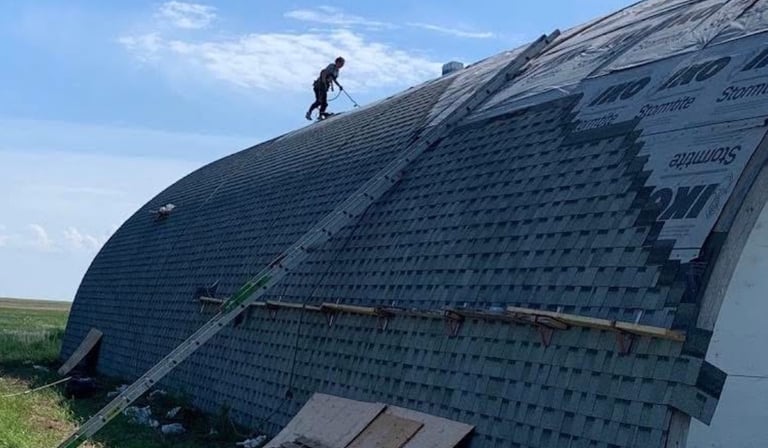 Professional roofers installing IKO shingles on a large curved barn roof using safety harnesses.