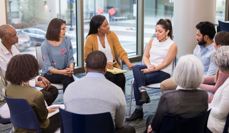 a group of people sitting in a circle with a woman in a white shirt