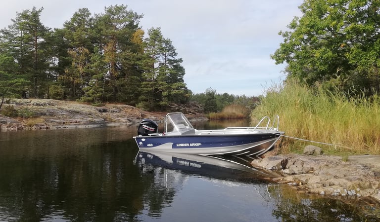 Auf einem See in Schweden liegt das Boot „Linder Arkip“. Es gibt Kiefern und Felsen.