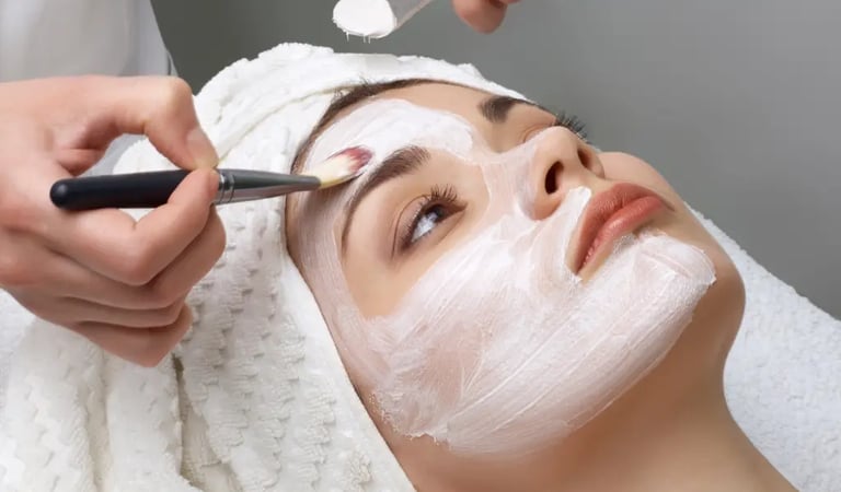A woman with a white towel on her head receiving a professional facial treatment with a  brush