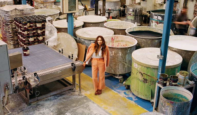 A woman in a coral suit standing in a professional paint factory with industrial mixing vats and conveyors.
