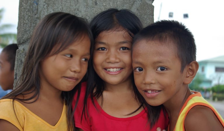 Three smiling Filipino children posing closely together outdoors in a rural village setting.