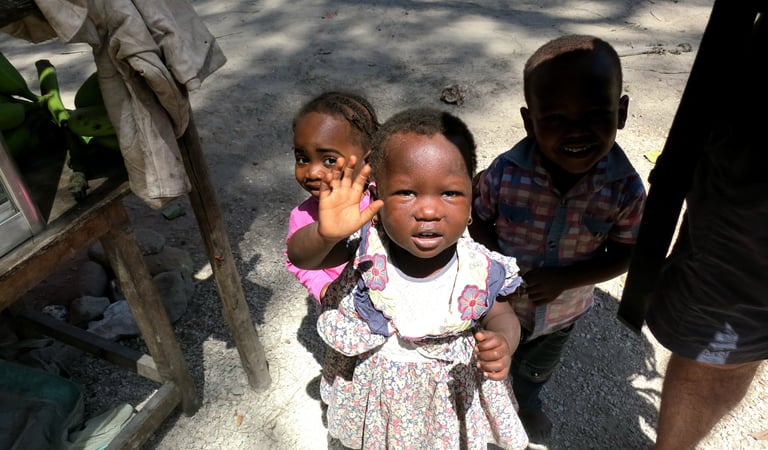 Three young African children standing outside and smiling, with one girl waving her hand.