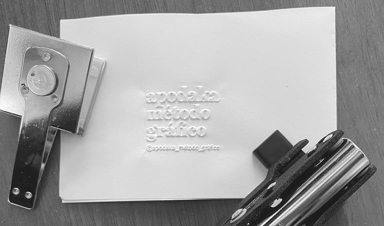 Sello metálico estampando en relieve el logotipo 'apodaka método gráfico' sobre papel blanco