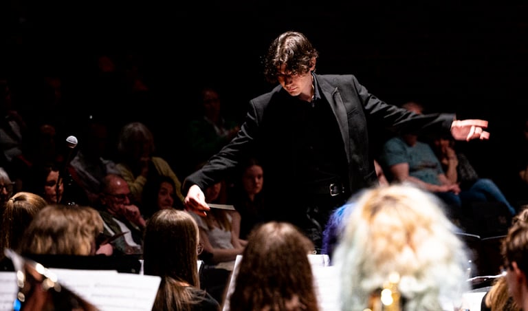 Conductor in black, with arms spread, amid heads of orchestra-members.