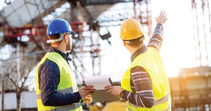 Two construction engineers wearing hard hats and safety vests inspect a building site.