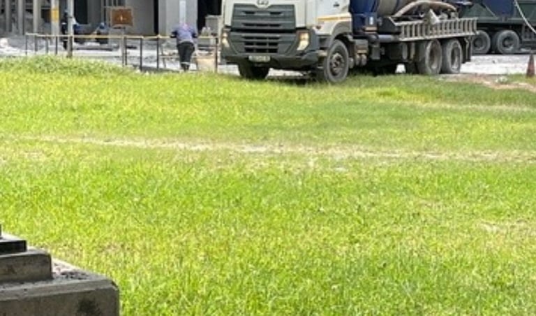 A white industrial vacuum truck parked outside a large concrete factory facility on a sunny day.