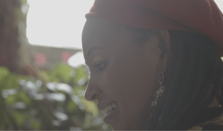 A person wearing a beanie with medium brown skin and black dreads smiling and looking down.