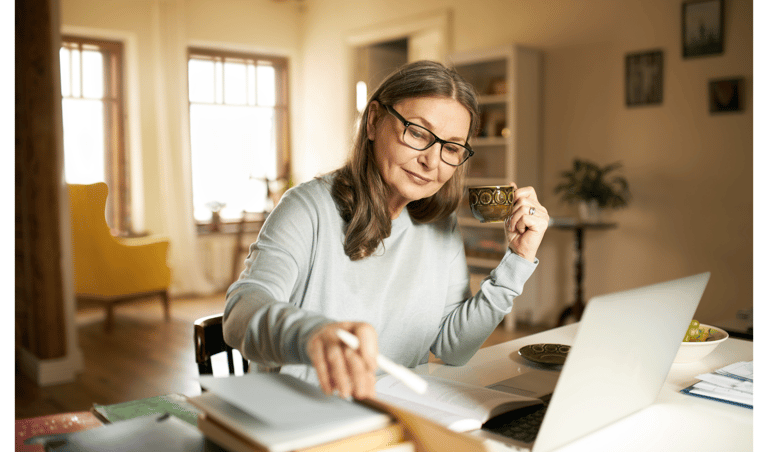 A senior woman wearing glasses drinks tea while working on a laptop at her home office desk.