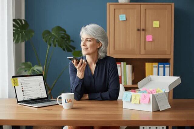 Woman at desk recording a voice note, surrounded by sticky notes and an open laptop. LVA mug visible