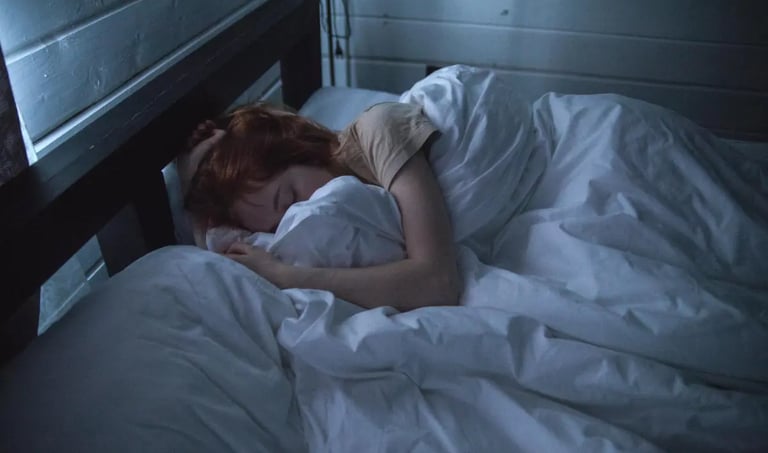 A redhead woman sleeping deeply in a dark bedroom with white bedding and a wooden bed frame.