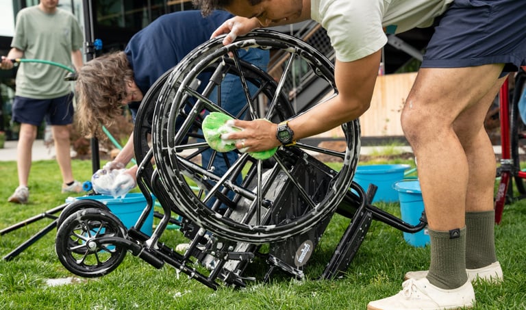A wheelchair flipped upside down on the grass with someone sponging the wheels