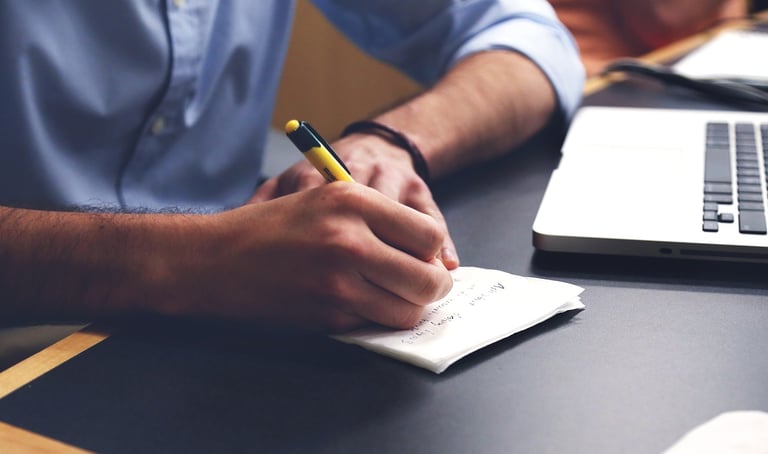 A person writing notes with a yellow pen on paper next to a laptop on a desk.